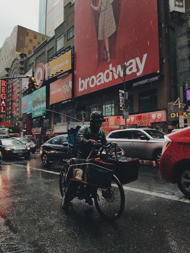 Bicycle courier in New York City with delivery bags, riding past ads and storefronts alongside traffic on 8th Avenue during a rainy day