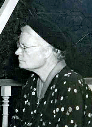 Black-and-white photo of Dorothy Day in profile, wearing dark cap and glasses, 1950s