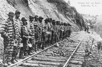 Black and white photo of men wearing white and black striped prison uniforms working on a chain gang in 1915
