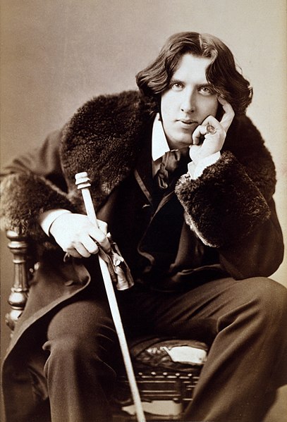 Photographic portrait of Oscar Wilde, sitting in chair dressed in suit and coat with fur collar