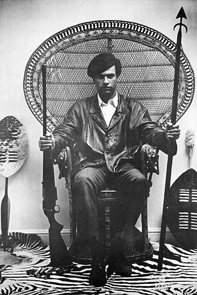 Portrait of Huey Newton sitting in a rattan chair with a rifle and a spear