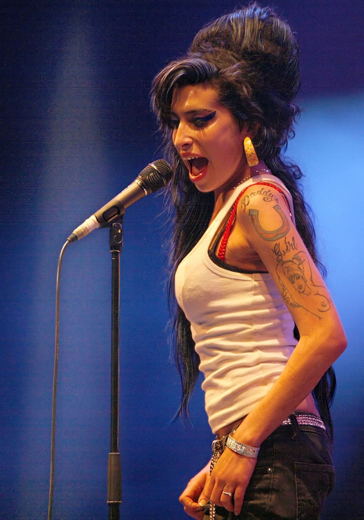 Amy Winehouse with her signature beehive wearing a white tanktop and singing into a microphone