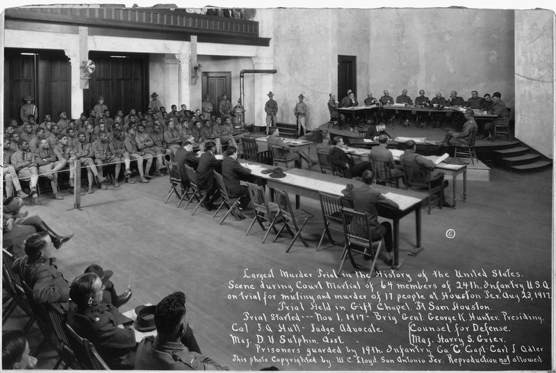 Black-and-white photograph of court martial trial for Camp Logan Mutiny, showing accused soldiers seated in roped off section on side of the room and others seated at tables in center and back