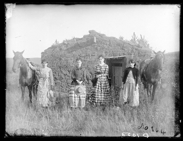 Black and white photo from 1886 of four women and two horses standing in front of a shack