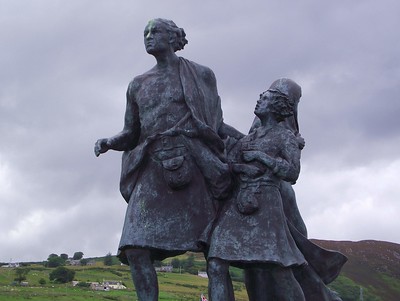 Photo of the Emigrants statue, commemorating Highlands and Islands people who left Scotland in the Clearances and depicting a kilted father and son advancing while the mother looks back, with dark cloudy sky and green slopes visible in background