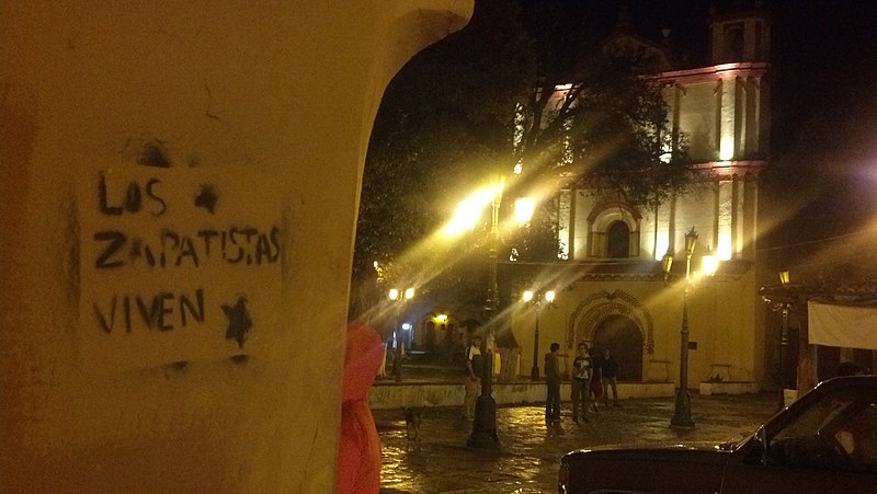 Nighttime photograph of graffiti on a wall reading 'LOS ZAPATISTAS VIVEN' (The Zapatistas live) in black all-caps lettering, with lights in courtyard visible in background to the right