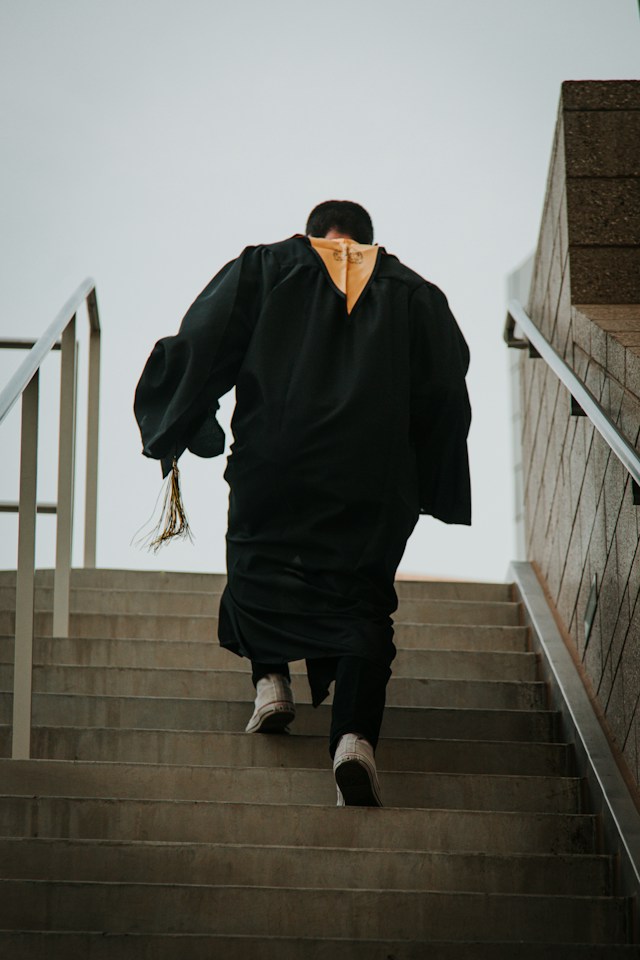 Graduate in gown walking up steps with cap in hand, facing away from the camera