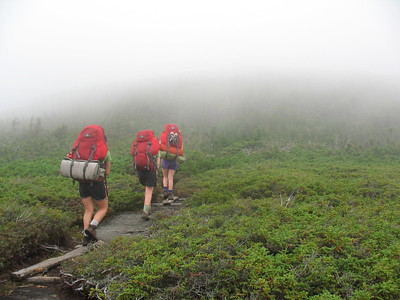 Photo of three hikers carrying red packs and facing away from camera while ascending a slope on the Appalachian Trail into mist ahead