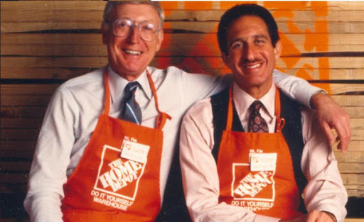 Home Depot founders Marcus and Blank wearing orange Home Depot aprons posed in front of lumber