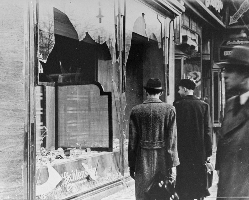 Two men stand in front of a shattered storefront window after Kristallnacht