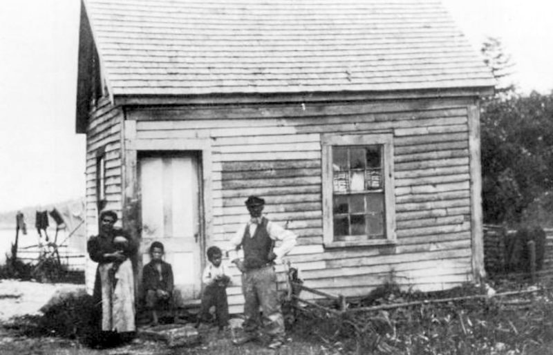 Black and white photo of dark-skinned family standing in front of a small house on Malaga Island