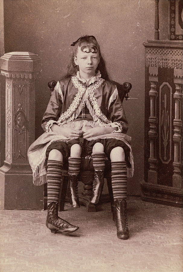 Black and white photo of Myrtle Corbin as a girl in ornate clothing sitting on a chair with her four legs visible