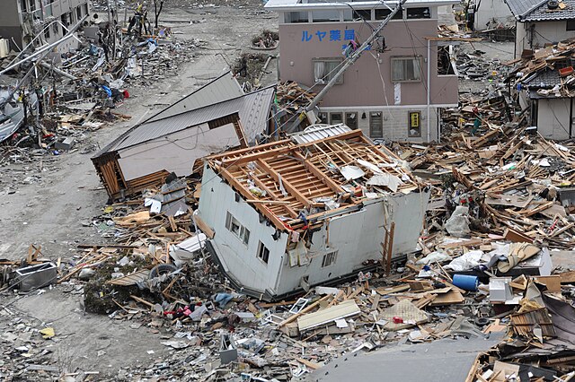 Color photo of upended house, debris and surrounding damage caused by Tohoku earthquake and tsunami
