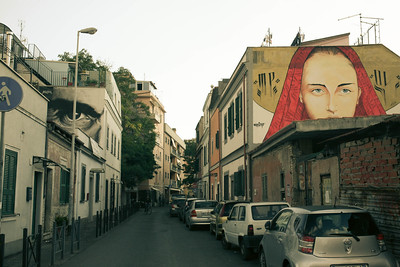 Street view of the Pigneto neighborhood of Rome, location of the Barikamà warehouse, showing parked cars and detailed art depicting human faces on buildings