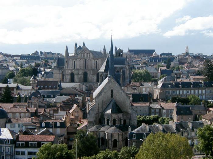 Panoramic view of Poitiers showing a Gothic cathedral rising above a cluster of medieval stone buildings with red‑tiled roofs, surrounded by greenery under a partly cloudy sky