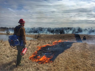 Photo of a controlled burn at the Froland Waterfowl Production Area in Minnesota, showing a worker wearing fire protection gear and standing in front of a burning strip of prairie