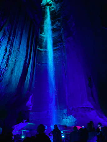 A dark cave featuring a waterfall lit by a bright blue light with visitors in the foreground taking pictures