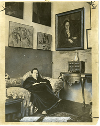 Black and white photo of Gertrude Stein sitting on a couch with her portrait hanging on the wall to the right side