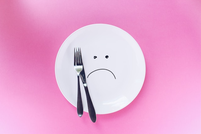 Sad face drawing on empty white plate holding silverware against solid pink background