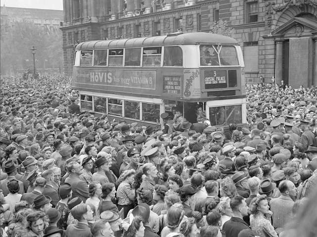 Black-and-white photo of V-E Day celebrations in London, England, showing a double-decker bus in the middle of the street surrounded by a crowd, with Westminster Abbey visible in background