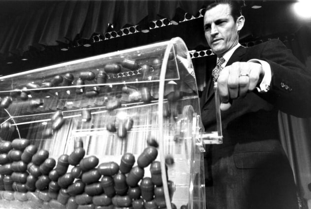 A man in a suit turns the crank on a glass lottery machine holding the capsules containing birthdays for the Vietnam War draft