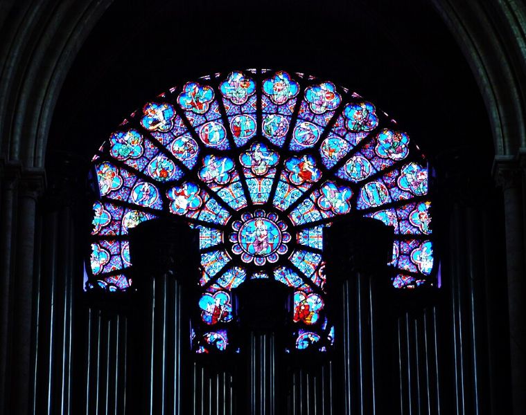 Circular stained-glass window partly obscured at the bottom by the silhouettes of organ pipes, similar to the North and South windows but with more image detail visible, including human figures