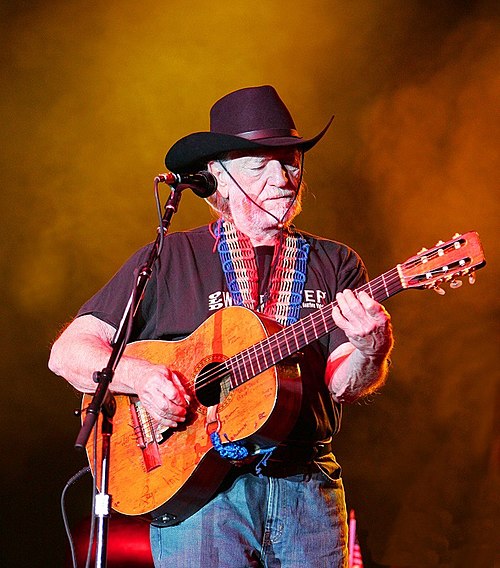 Willie Nelson, an older bearded white man, playing guitar and wearing a cowboy hat