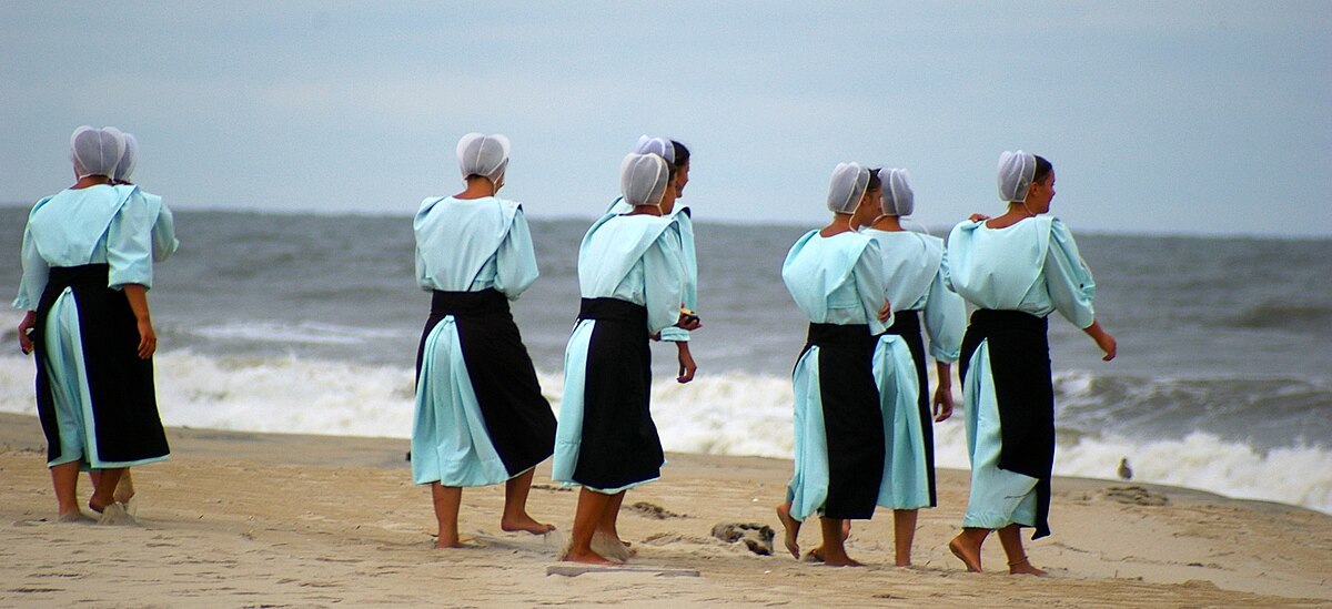 A group of young women in identical dresses and bonnets standing barefoot in sand near the water